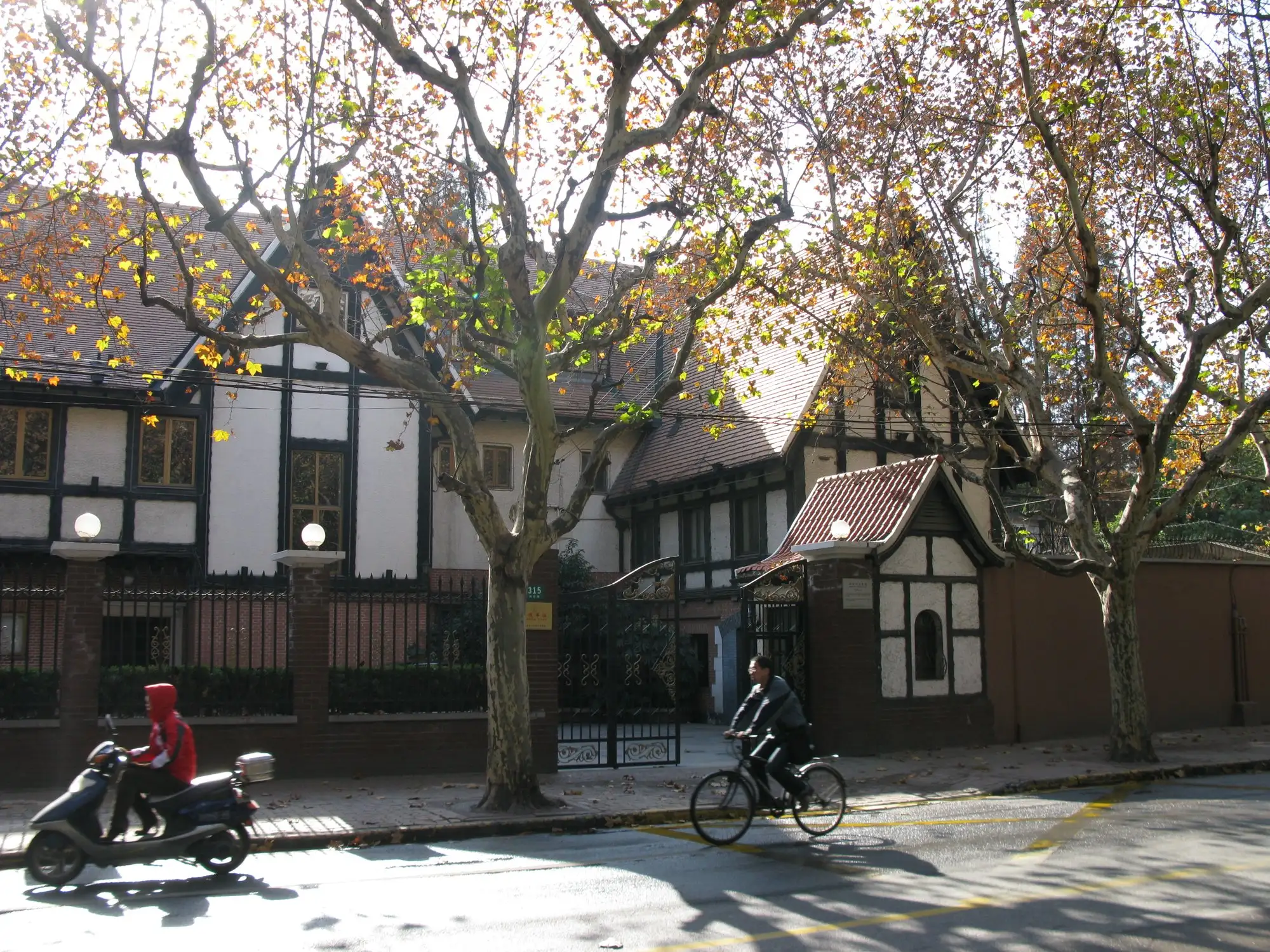 Half-timbered villa with steep tiled gables behind a brick and iron fence, plane trees with autumn leaves, a cyclist and a scooter on the street in front.