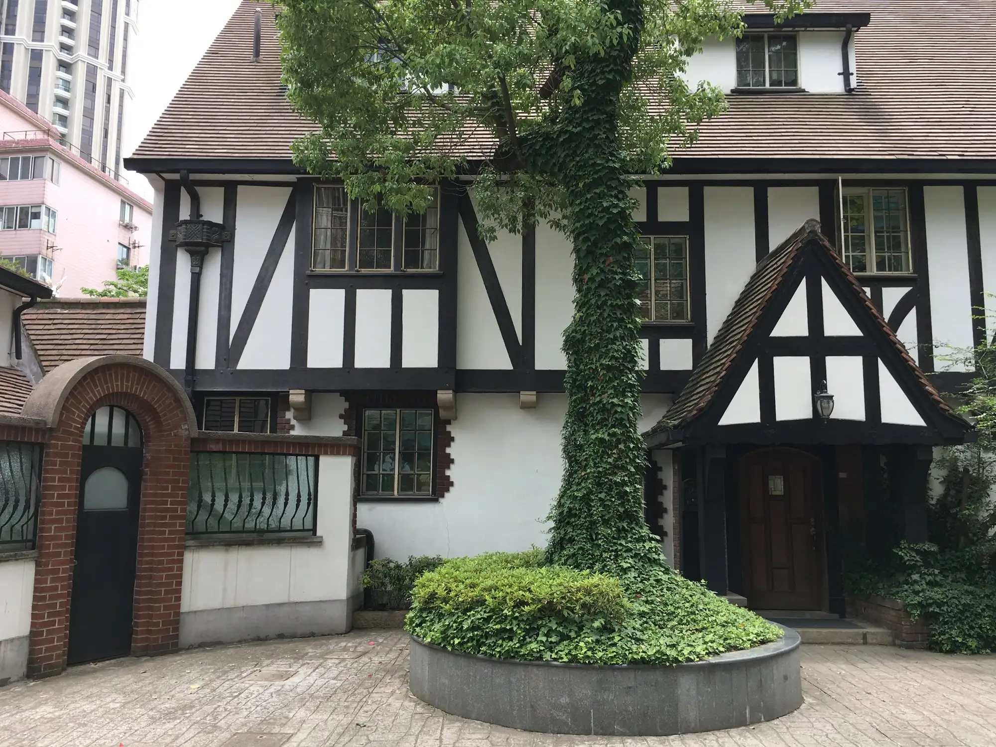 Black-and-white half-timbered house with a steep brown roof and a small gabled porch, a large ivy-covered tree in a round planter in front.