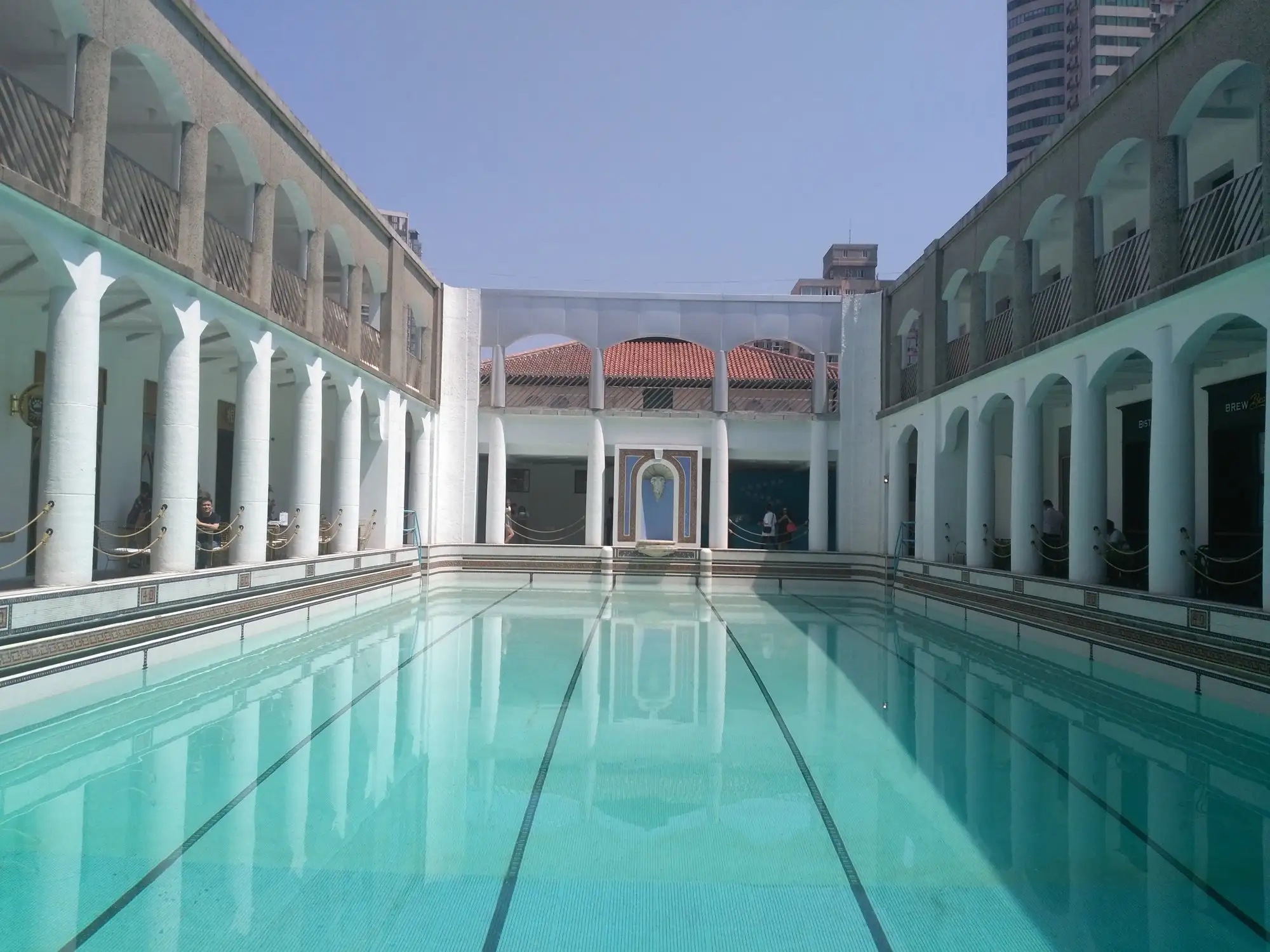 Long rectangular swimming pool of pale blue water framed by white arcades on two sides and a red-tiled pavilion at the far end.
