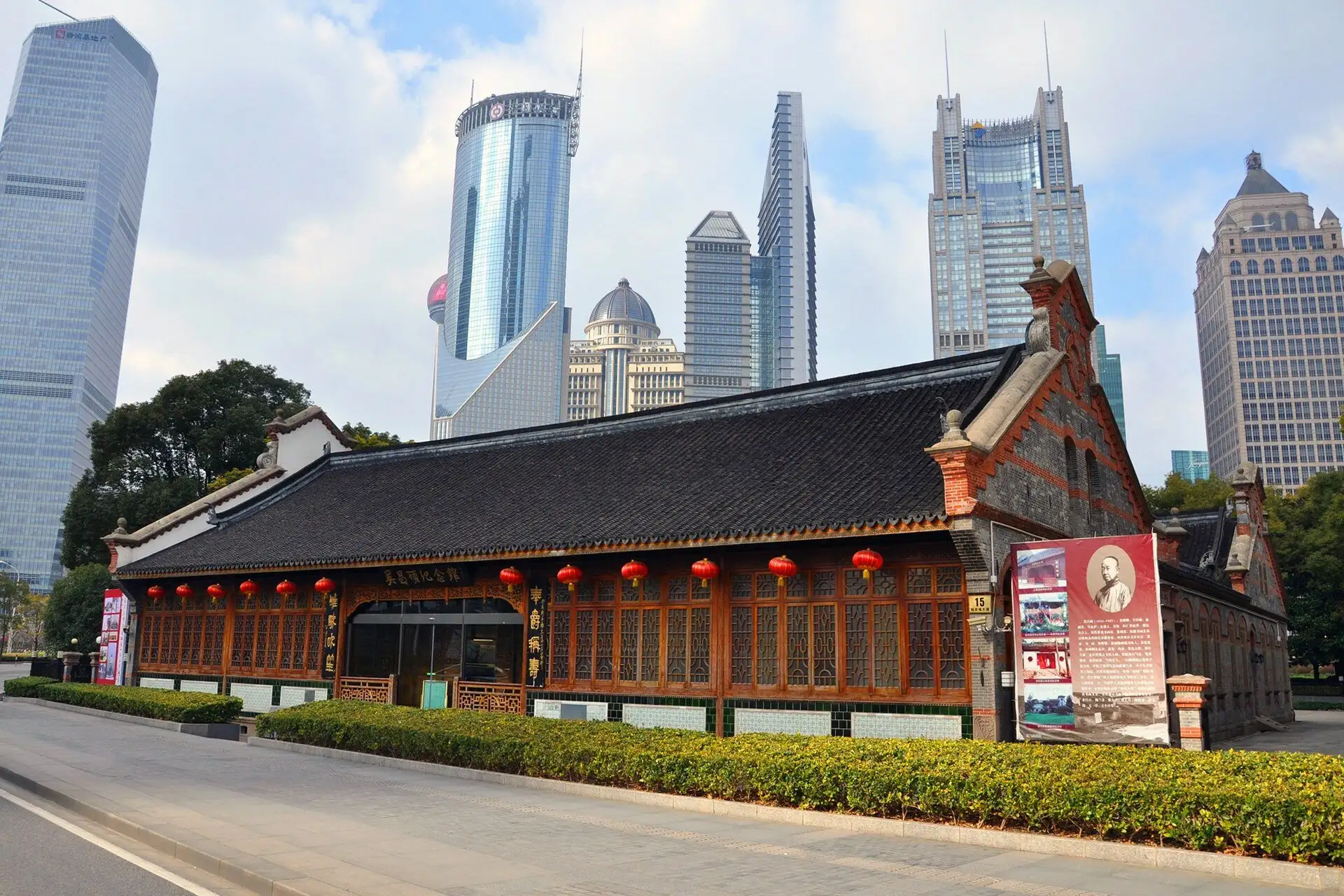Long low building with a dark tiled Chinese roof, carved wooden windows hung with red lanterns and a stepped red-and-grey brick gable at one end, in front of glass skyscrapers.