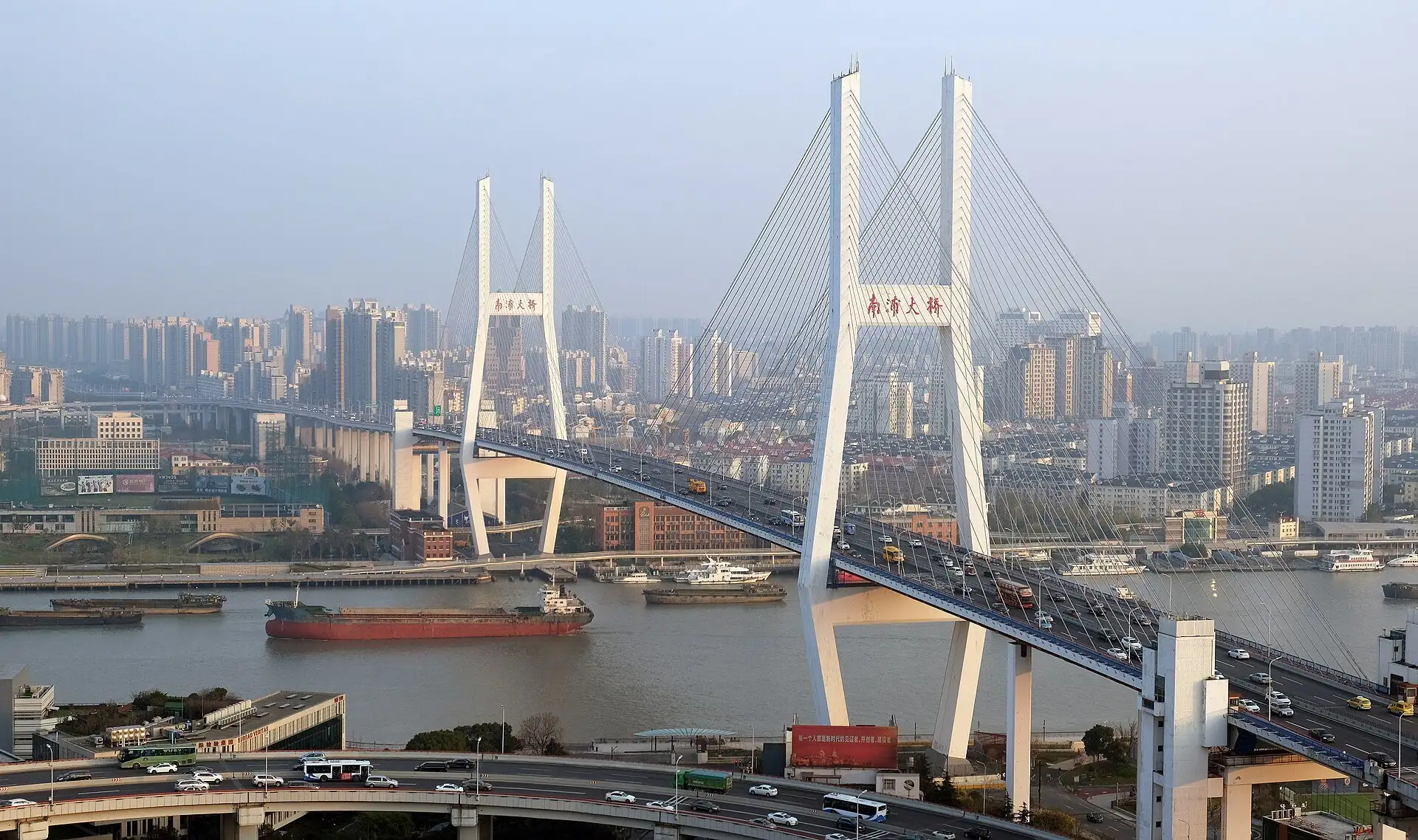 White cable-stayed bridge with two A-shaped towers carrying traffic across a river, a red cargo ship passing beneath and apartment blocks on the far bank.