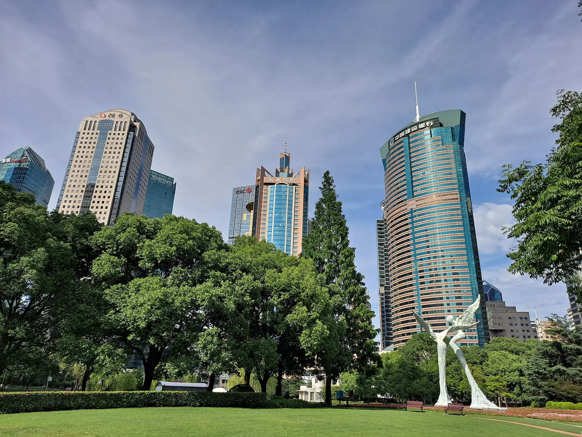 Wide green lawn with large trees and a white winged sculpture, surrounded by office towers under a blue sky.