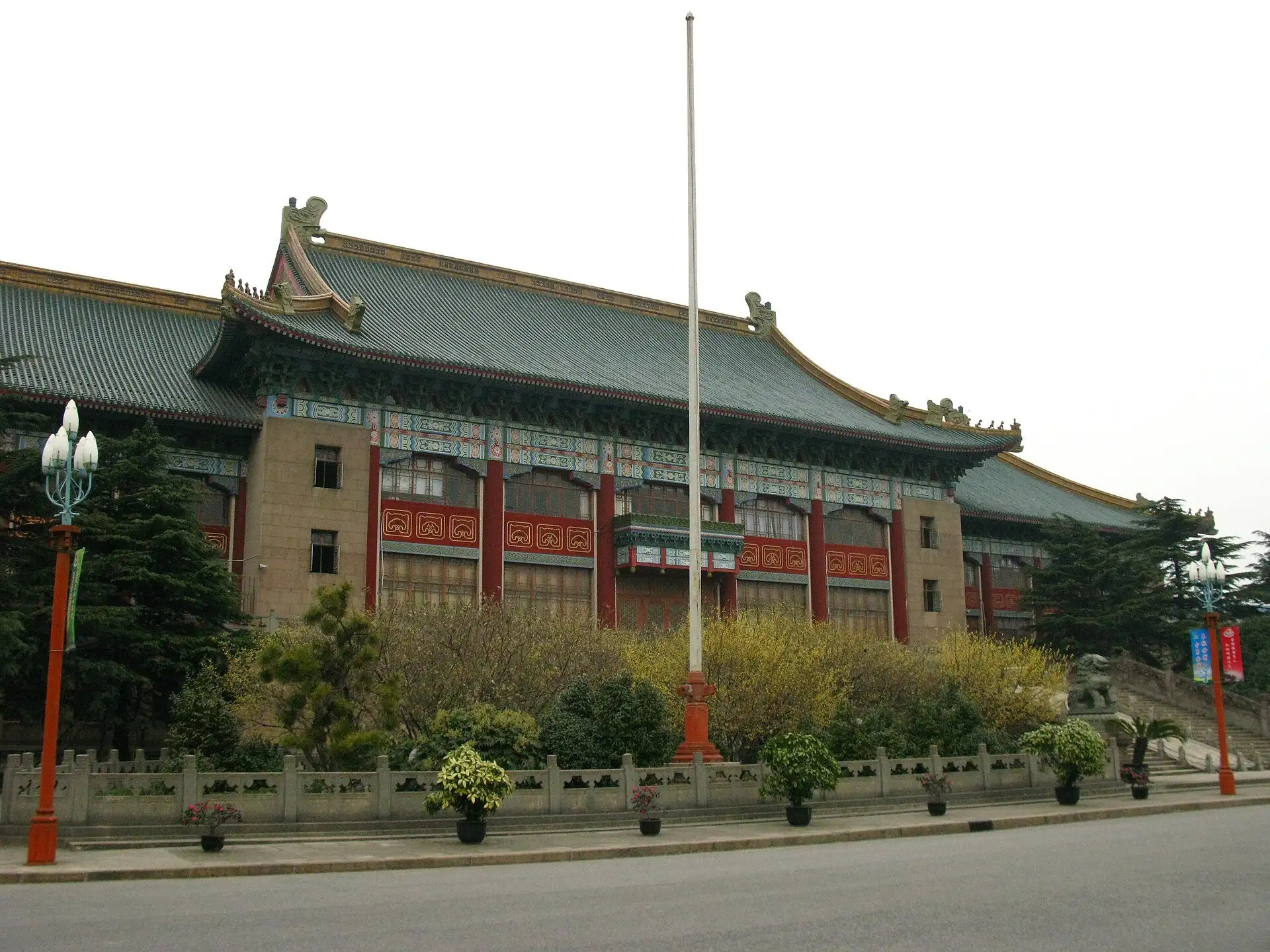 Large building with a green-tiled hipped roof with upturned ridges, painted brackets and red columns over a stone base, a flagpole and shrubs in front.