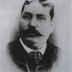 Black-and-white oval portrait of a heavy-set man with a dark moustache, wearing a checked tie and dark jacket.