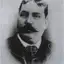 Black-and-white oval portrait of a heavy-set man with a dark moustache, wearing a checked tie and dark jacket.