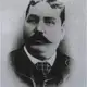 Black-and-white oval portrait of a heavy-set man with a dark moustache, wearing a checked tie and dark jacket.