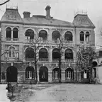 Black-and-white photograph of a three-storey brick-and-stone building with arched verandas, seen across a gravel courtyard.
