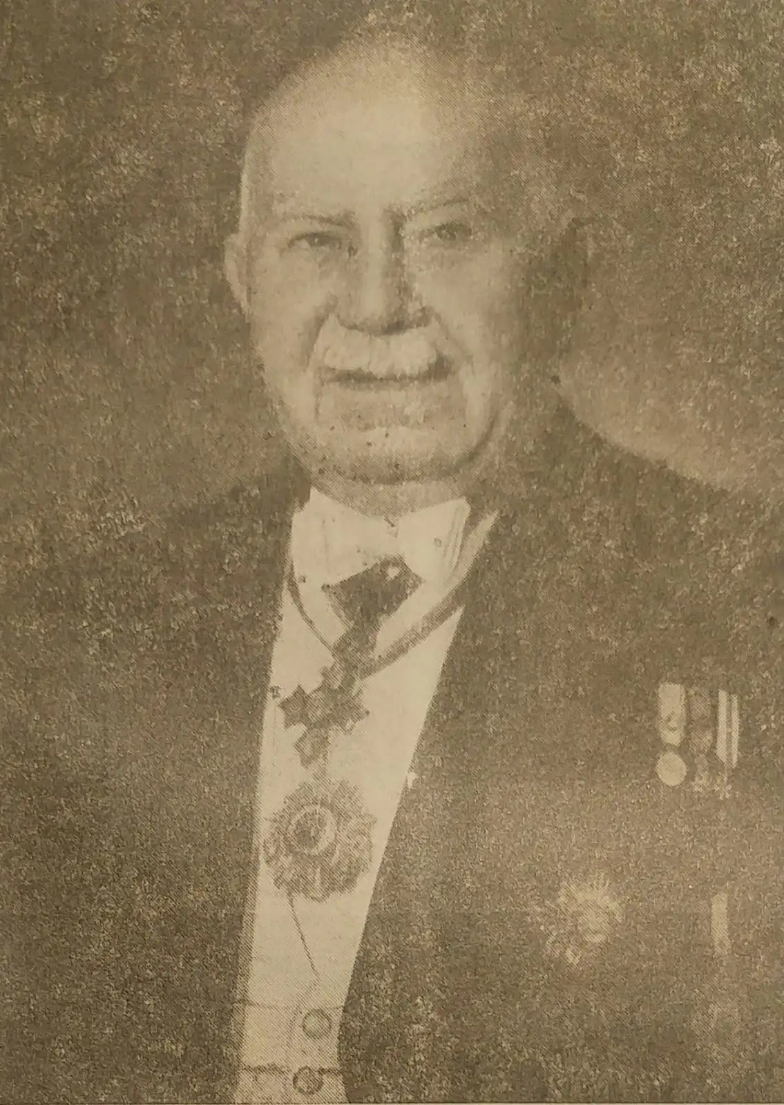 Faded newspaper-style photograph of an elderly man with a white moustache in evening dress with a sash and medals.