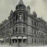 Black-and-white photograph of a corner building of red brick and stone with a conical tower, balconies and bamboo scaffolding on one side.