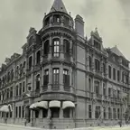 Black-and-white photograph of a corner building of red brick and stone with a conical tower, balconies and bamboo scaffolding on one side.