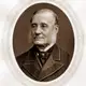 Sepia oval portrait of an elderly man with white side-whiskers in a dark coat with a velvet collar.