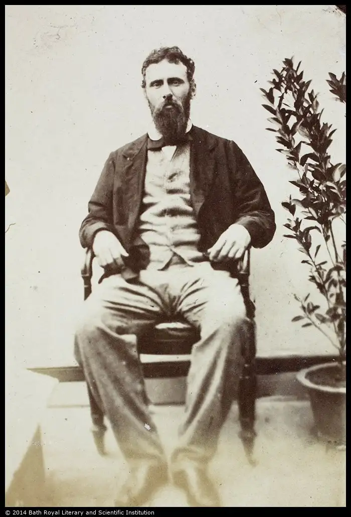Sepia photograph of a bearded man in a dark jacket and bow tie, seated on a chair beside a potted plant.