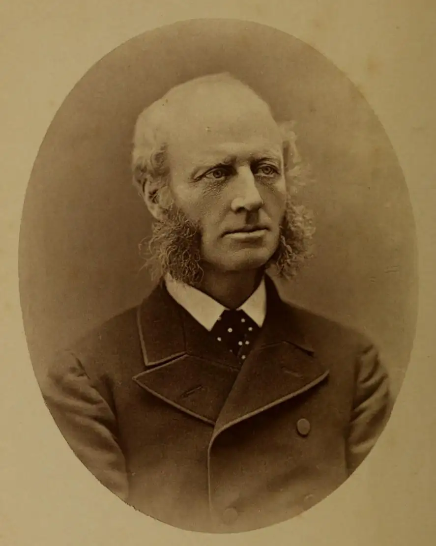Sepia oval studio portrait of a middle-aged man with a bald head and long curly side-whiskers, in a dark coat.