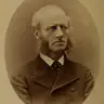 Sepia oval studio portrait of a middle-aged man with a bald head and long curly side-whiskers, in a dark coat.