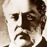 Sepia close portrait of a man with swept hair and a grey beard, in a dark coat.