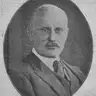 Oval black-and-white portrait of a balding man with a moustache in a suit and tie.