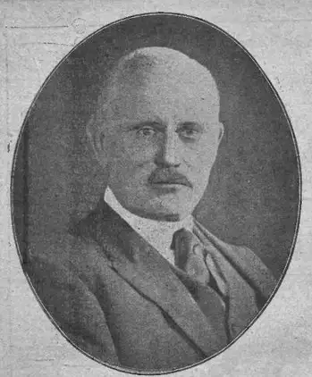 Oval black-and-white portrait of a balding man with a moustache in a suit and tie.