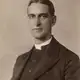 Sepia studio portrait of a clean-shaven man in a dark clerical jacket with a white collar.