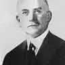 Black-and-white portrait of a balding man with a small moustache in a dark suit and tie.