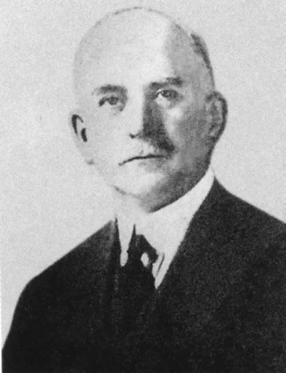 Black-and-white portrait of a balding man with a small moustache in a dark suit and tie.