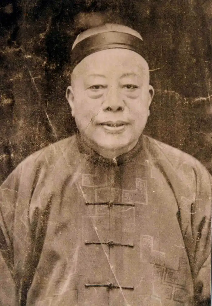 Sepia portrait of an older man in a small cap and a patterned silk jacket, on a cracked and stained print.