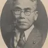 Black-and-white oval portrait of an elderly man in round glasses, jacket and tie, with a small pin on the lapel.
