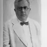 Black-and-white portrait of a man in round glasses, a white jacket and a bow tie, with a printed caption below.