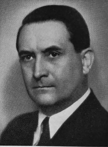 Black-and-white portrait of a dark-haired man in a dark suit and tie looking toward the camera.