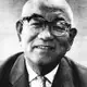 Black-and-white close portrait of a smiling bald elderly man in thick-rimmed glasses, jacket and patterned tie.