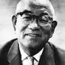 Black-and-white close portrait of a smiling bald elderly man in thick-rimmed glasses, jacket and patterned tie.