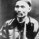 Black-and-white portrait of a man with a moustache wearing a skullcap and a dark silk jacket with toggle buttons.