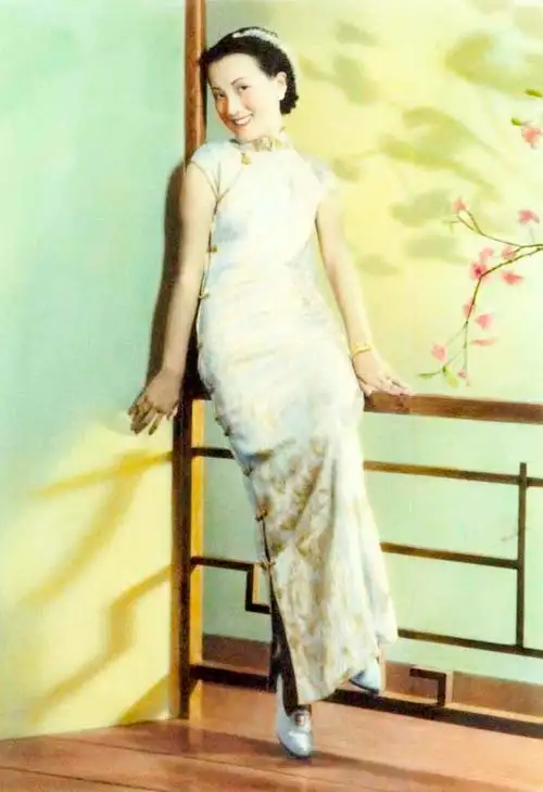 Hand-tinted photograph of a smiling young woman in a long pale qipao leaning against a wooden railing, a branch of pink blossom painted on the wall beside her.