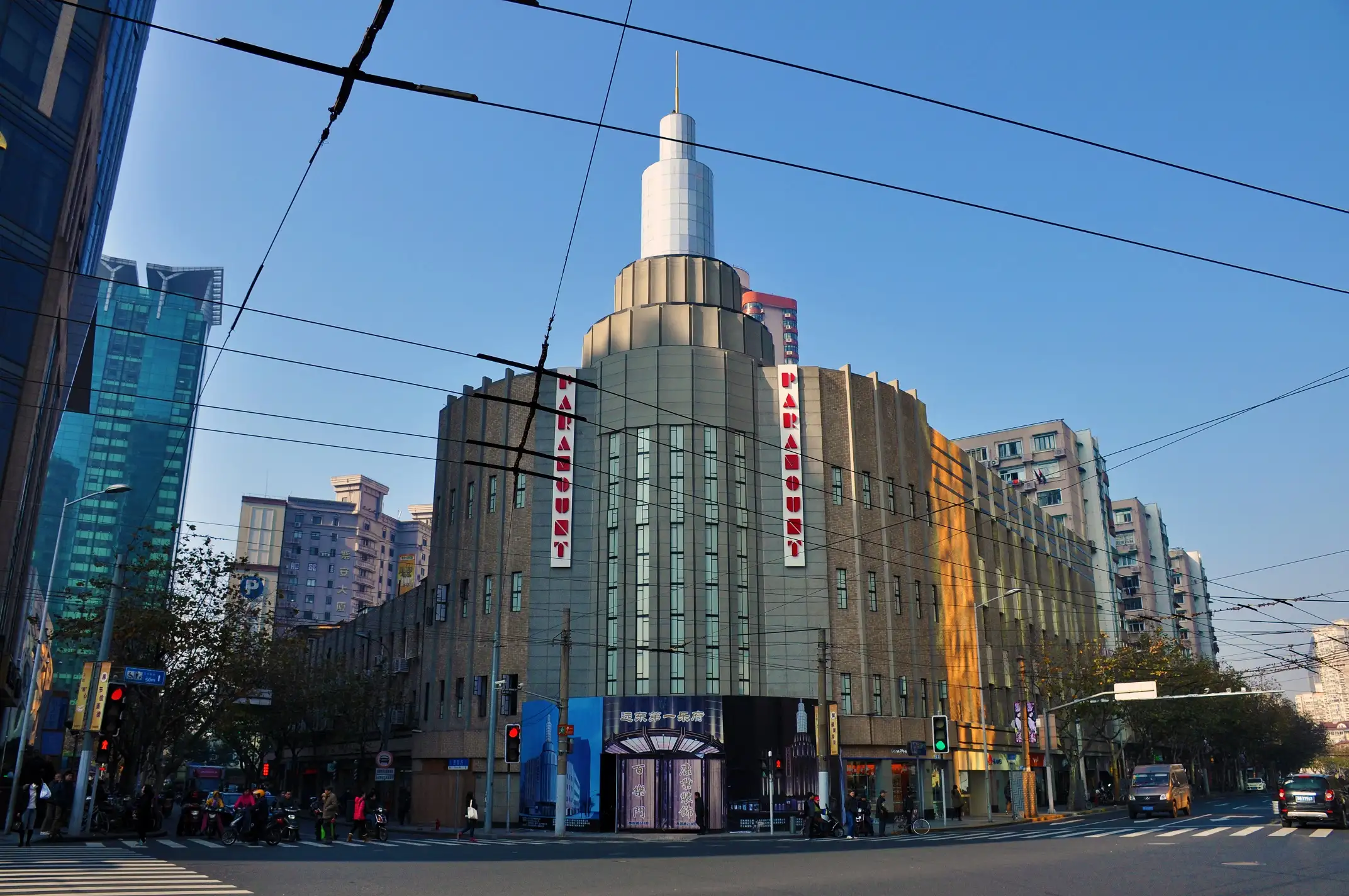 Grey Art Deco corner building with a round stepped tower rising above the entrance, two vertical red PARAMOUNT signs, trolleybus wires criss-crossing a blue sky and pedestrians at the crossing.