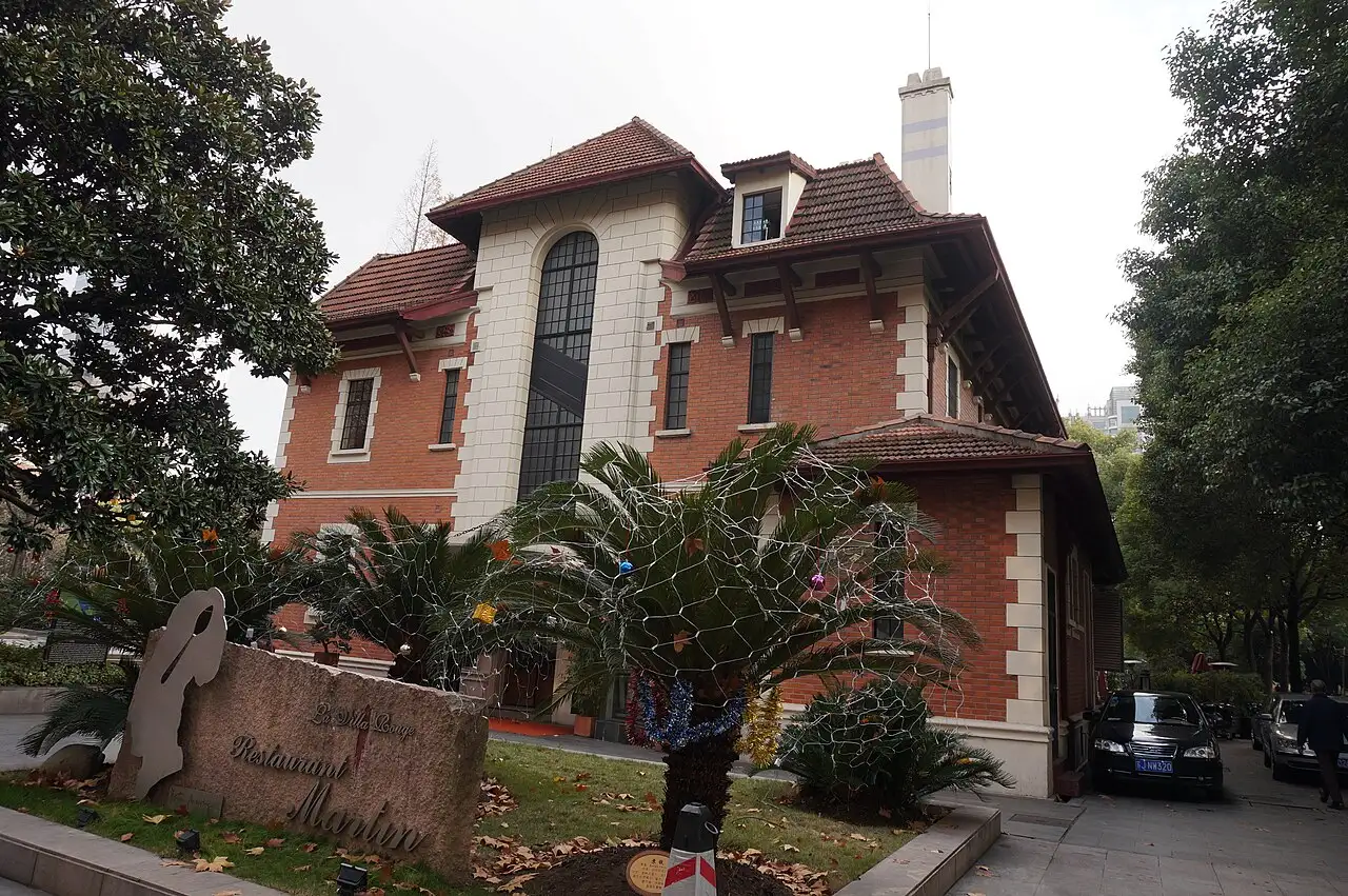 Two-storey red-brick house with white stone quoins, a tall arched staircase window and deep tiled eaves, palms and a magnolia in front, a carved stone sign at the lower left.