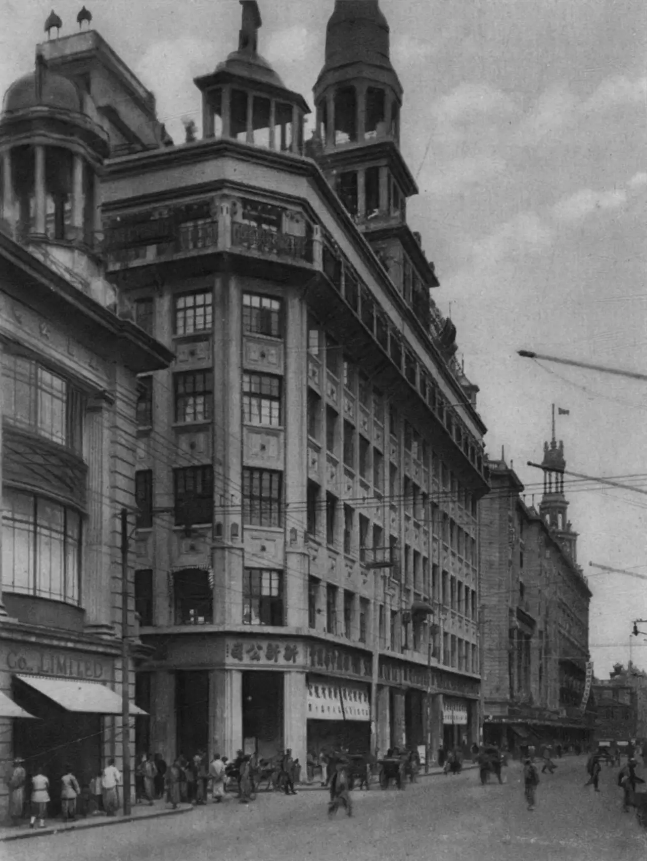 Black-and-white photograph of a plain seven-storey corner building with a Chinese signboard over the ground floor, open-sided pavilions and a spire on the roof, rickshaws and pedestrians in the street.