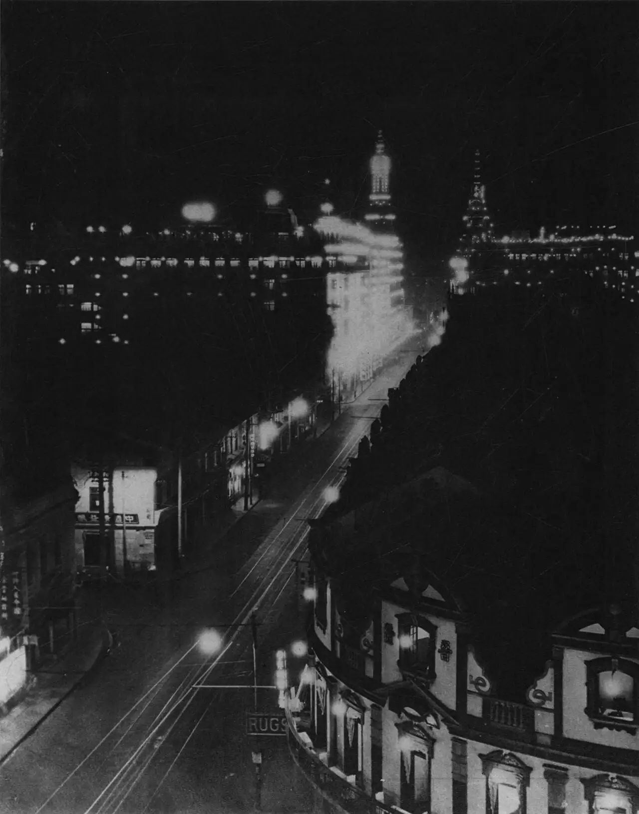 Night photograph looking down a street lined with lit windows, two towers in the distance outlined with rows of light bulbs, streaks of light from passing traffic.