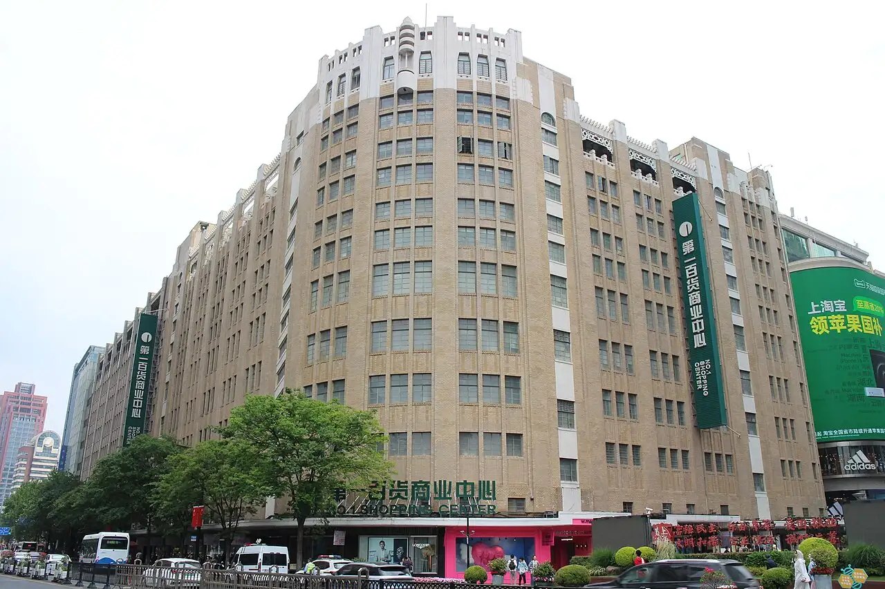 Ten-storey buff-brick building with a rounded corner and vertical window bands, green vertical signs reading 第一百货商业中心, trees and traffic in front.